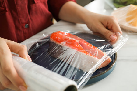 Woman putting plastic food wrap over plate with salmon at white marble table in kitchen, closeupの写真素材