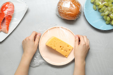 Woman putting plastic food wrap over plate with cheese at gray table, top view