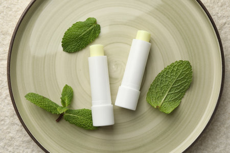 Mint lip balms and green leaves on beige textured table, top view. Cosmetic product