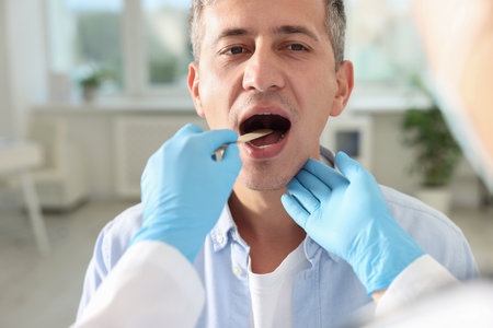 Doctor examining man's throat with tongue depressor in clinicの写真素材