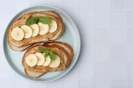 Tasty sandwiches with peanut butter, banana and mint on white tiled table, top view. Space for textの写真素材
