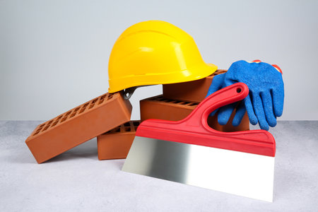 Red bricks, plastering trowel, gloves and hardhat on textured table against light background