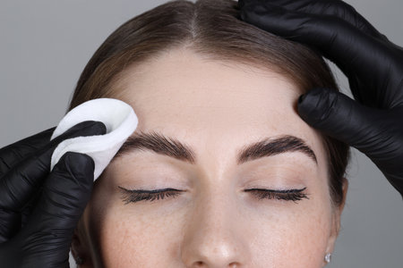Brow lamination. Cosmetologist wiping woman's eyebrows with cotton pad against gray background, closeupの写真素材