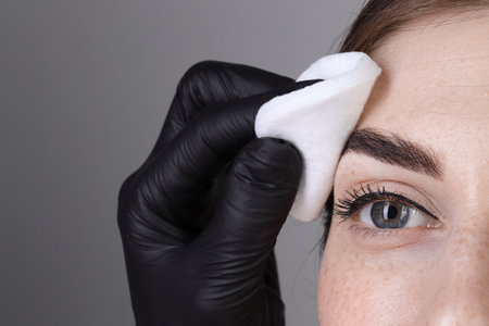 Brow lamination. Cosmetologist wiping woman's eyebrows with cotton pad against gray background, closeupの写真素材
