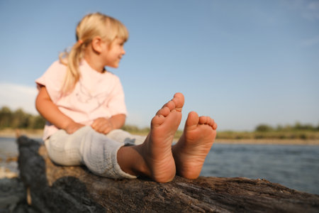 Cute little girl sitting on tree trunk near river, selective focus. Child enjoying beautiful natureの写真素材