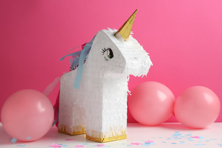 Bright pinata in shape of unicorn and balloons on white table against pink backgroundの写真素材