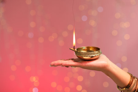 Diwali celebration. Woman holding lit diya lamp on color background with blurred lights, closeup. Space for textの写真素材