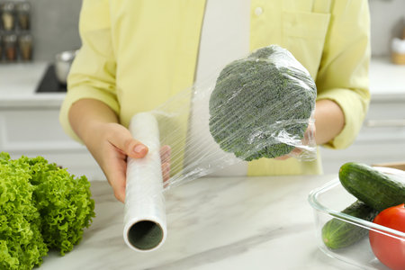 Woman putting plastic food wrap over broccoli at countertop in kitchen, closeupの写真素材