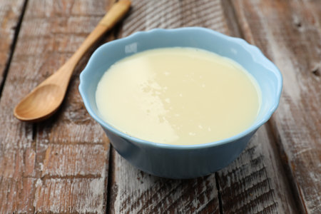 Condensed milk in bowl and spoon on wooden table, closeupの写真素材