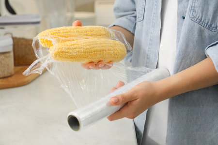 Woman putting plastic food wrap over corncobs at countertop in kitchen, closeupの写真素材