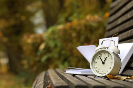 Autumn time. Alarm clock and book on bench in park, closeup. Space for textの写真素材