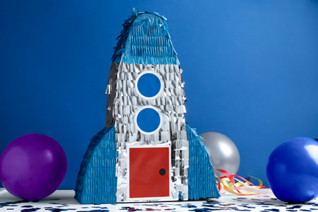 Bright pinata in shape of spaceship, balloons and confetti on table against blue background