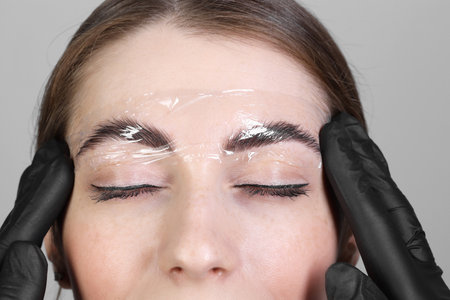 Brow lamination. Cosmetologist putting film onto woman's eyebrows against grey background, closeupの写真素材