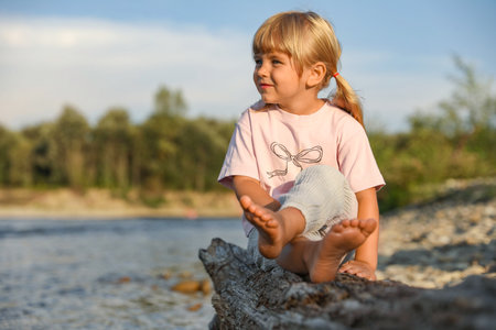 Cute little girl sitting on tree trunk near river. Child enjoying beautiful natureの写真素材