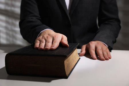 Man taking oath with his hand on Bible at white table, closeup