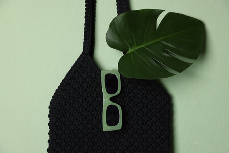 Handmade macrame shopping bag with leaf and sunglasses on light green background, top view