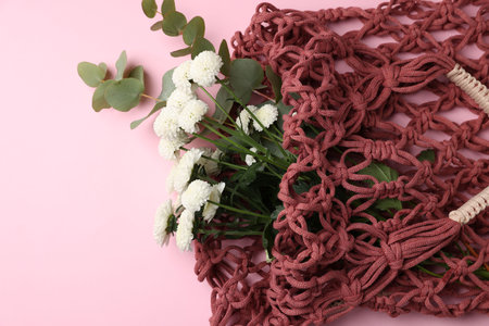 Macrame shopping bag with flowers and eucalyptus branches on pink background, above view