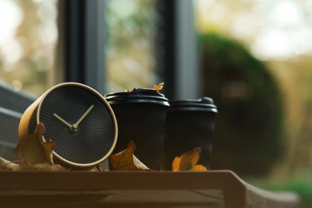 Autumn time. Alarm clock, fallen leaves and cups with hot drinks near cafe outdoors, space for text