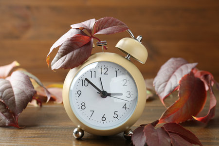 Autumn time. Alarm clock among leaves on wooden table, closeup