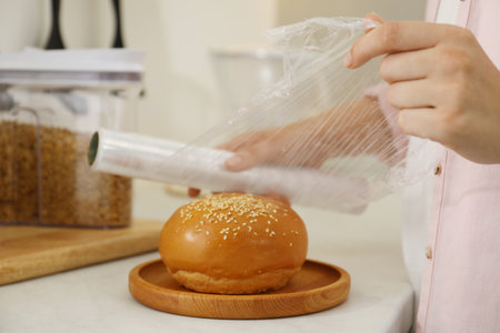 Woman putting plastic food wrap over fresh bun at white table in kitchen, closeupの写真素材