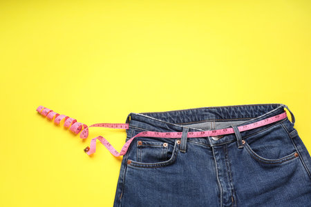 Jeans and measuring tape on yellow background, top view. Space for text