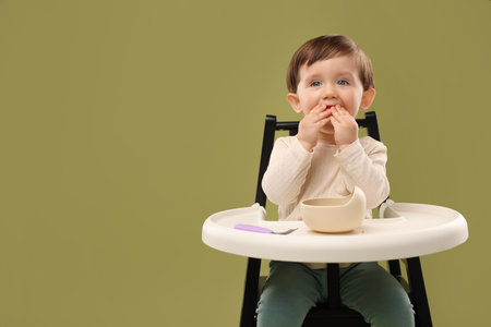 Cute little baby eating healthy food from bowl in high chair on olive background, space for textの写真素材