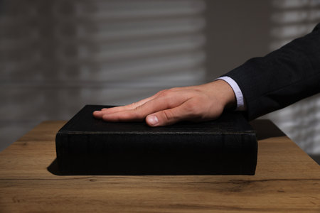 Man taking oath with his hand on Bible at wooden table, closeup