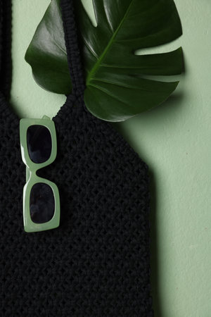 Handmade macrame shopping bag with leaf and sunglasses on light green background, top view