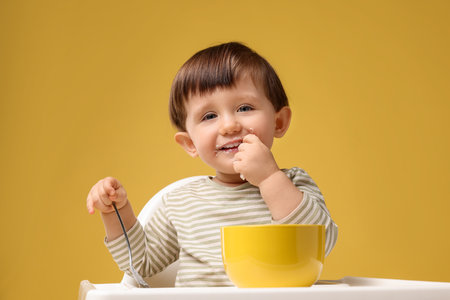 Cute little kid eating healthy baby food from bowl in high chair on yellow background