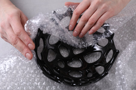 Woman covering ceramic bowl with bubble wrap at light gray table, closeup