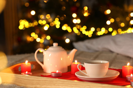 Aromatic tea in cup, teapot and burning candles on bed against blurred Christmas lights, closeup. Bokeh effectの写真素材