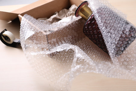 Beautiful glass vase in bubble wrap near cardboard box and adhesive tape on wooden table