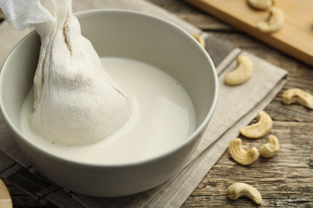 Making cashew milk. Cheesecloth and milk in bowl among nuts on wooden table, closeupの写真素材
