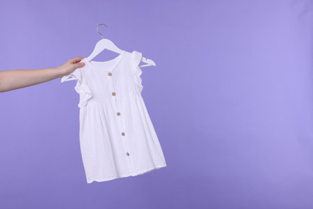 Woman holding hanger with white baby dress on light purple background, closeup. Space for textの写真素材