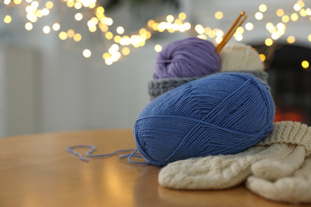 Colorful yarns, knitted mittens and crochet hooks on wooden table against blurred lights, closeup....