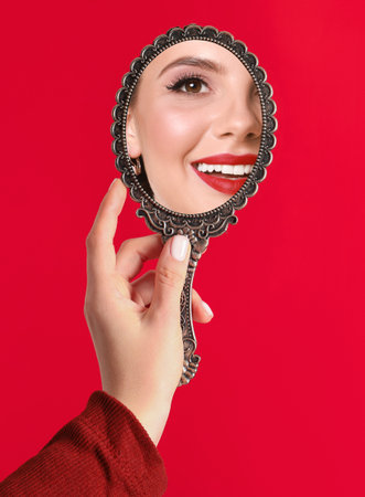 Beautiful woman looking in handheld mirror on red background, closeupの写真素材