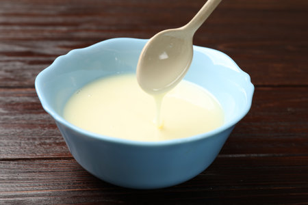 Condensed milk flowing down from spoon into bowl on wooden table, closeupの写真素材