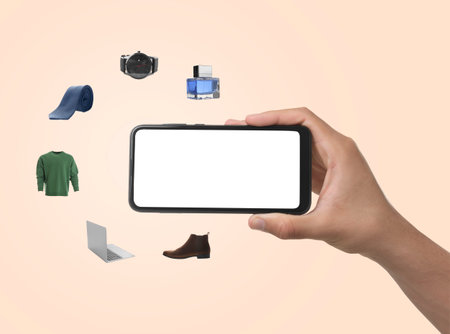 Man using mobile phone for online shopping on beige background, closeup. Different goods flying around. Device with blank screenの写真素材