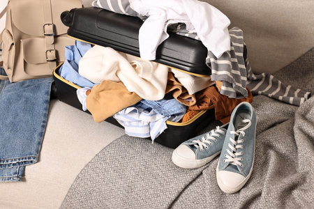 Open suitcase full of different messy clothes and sneakers on sofa indoorsの写真素材