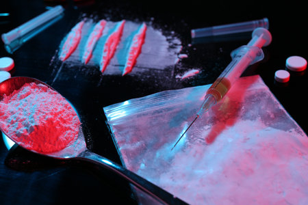 Drug addiction. Plastic bag with powder, syringe and spoon on black table in neon lights, closeup