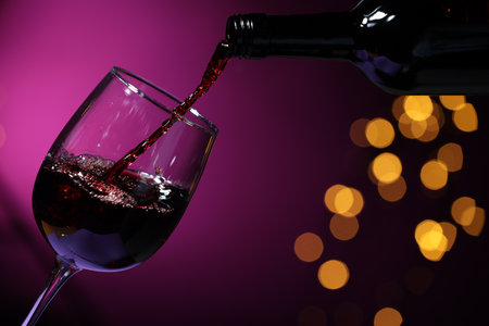 Pouring red wine into glass against purple background, closeup. Bokeh effect