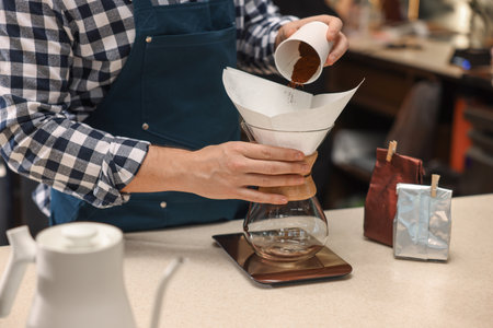Pouring ground coffee into glass coffeemaker with paper filter at table in cafe, closeup