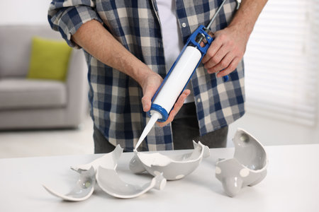 Using caulking gun to repair broken ceramic pieces at white table indoors, closeupの写真素材