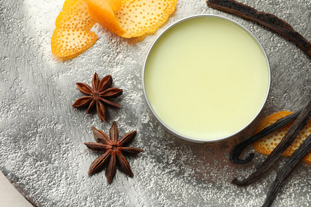 Natural solid perfume in container, vanilla pods, anise stars and orange peels on grey table, flat...