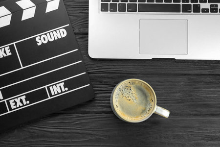Movie clapper, laptop and cup of coffee on black wooden table, flat lay