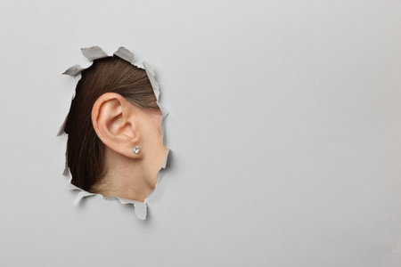 An ear visible through a torn hole in grey paper, closeup. Space for text