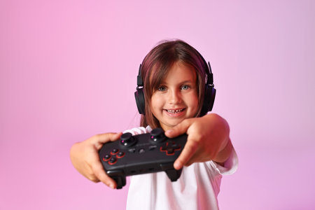 Happy little girl in headphones playing video game with controller on pink background