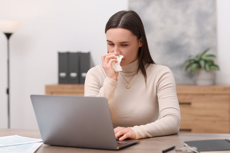 Sick woman with runny nose working at table in officeの写真素材