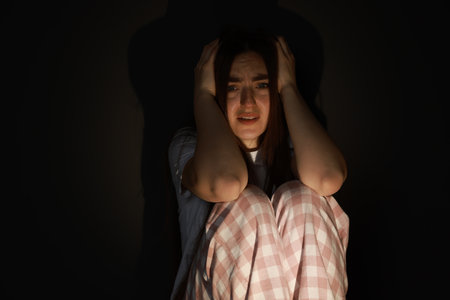 Fear of darkness. Scared young woman sitting on dark backgroundの写真素材