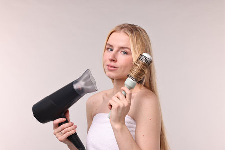 Beautiful young woman styling her hair with hairdryer and brush on light grey background